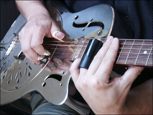 A musician playing slide guitar style. The slide is on his left ring finger. He is playing a National-type metal-body resonator guitar using fingerpicks on his right hand.